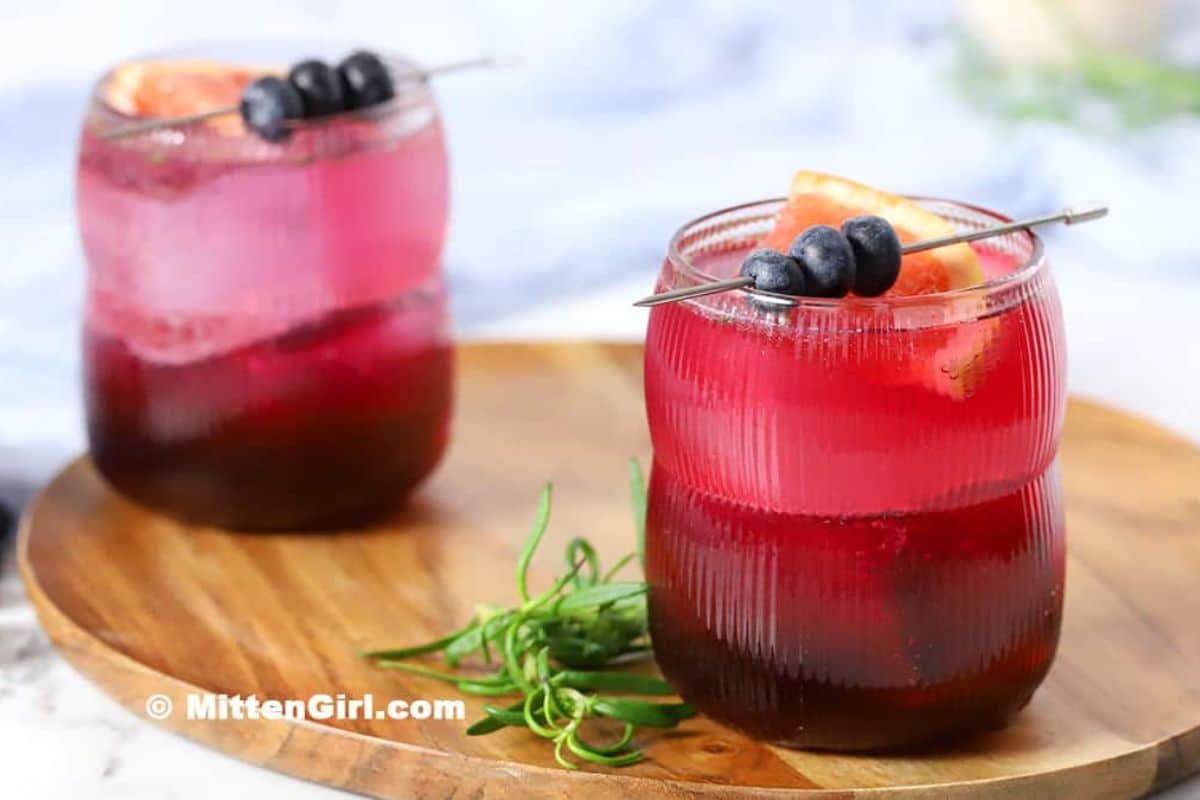 Two glasses of layered pink and dark red refreshing drinks, garnished with blueberries and orange slices, sit on a wooden tray with a sprig of greenery-perfect non alcoholic summer drinks for any gathering.