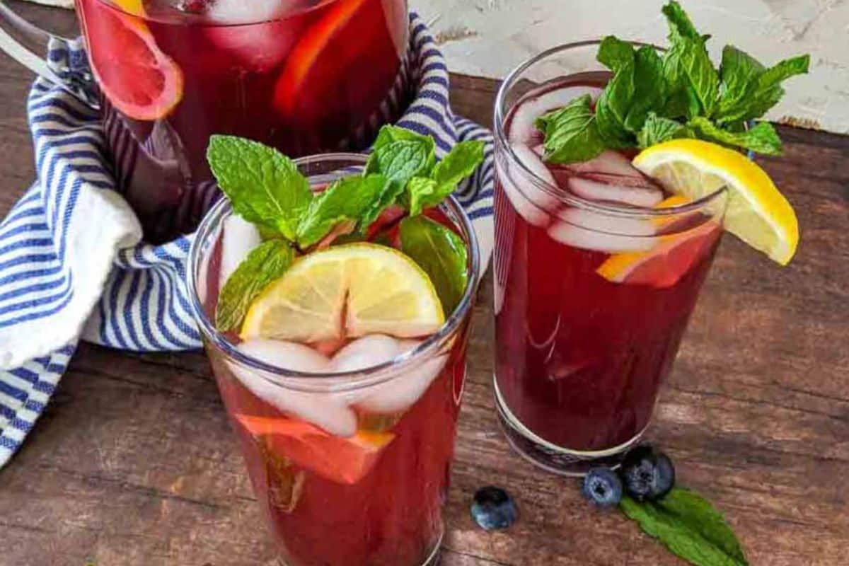 Two glasses and a pitcher of iced red fruit drink with lemon slices, mint sprigs, and blueberries on a wooden table—perfect for those seeking refreshing summer drinks or delicious non alcoholic summer drinks.