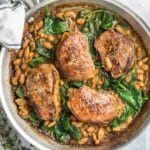 Four seared pork chops in a skillet with white beans and wilted spinach in a savory sauce.