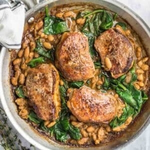 Four seared pork chops in a skillet with white beans and wilted spinach in a savory sauce.