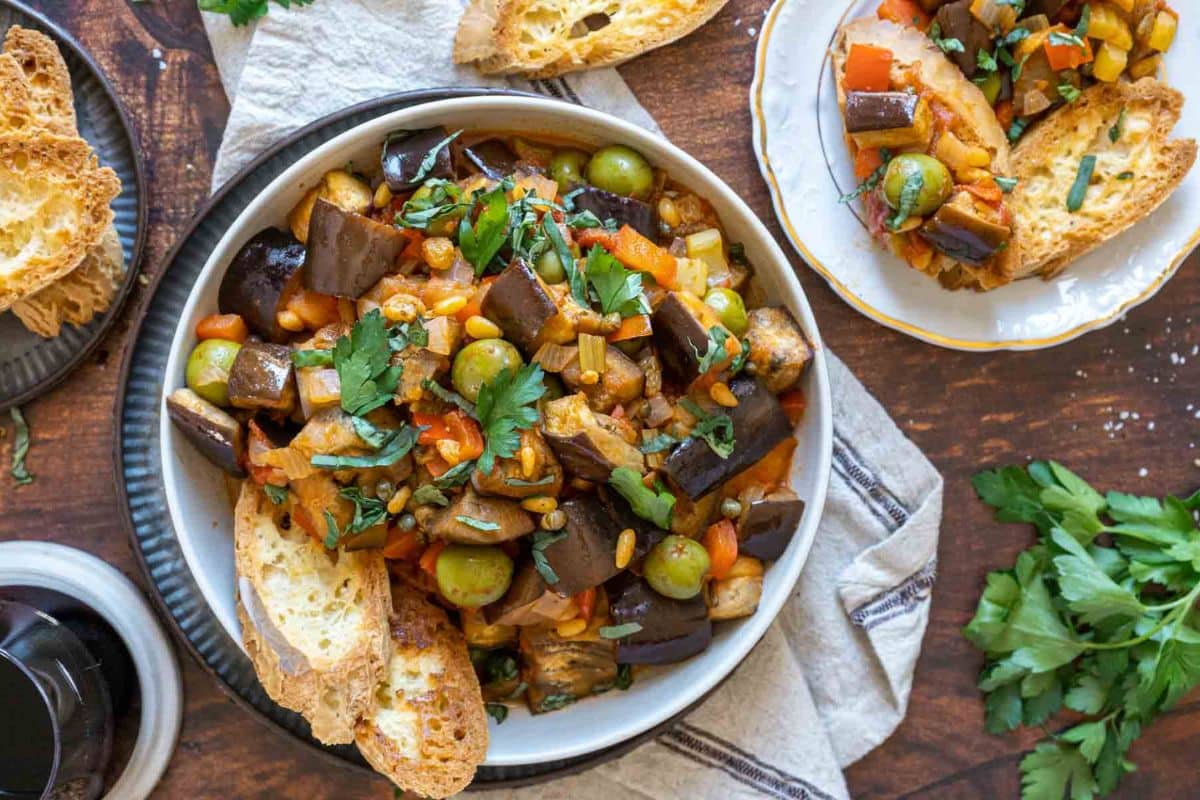A bowl of caponata with olives, eggplant, and herbs, served with toasted bread slices; a delicious example of eggplant recipes, set beside fresh parsley and a plate on a wooden table.