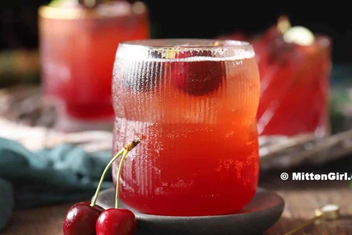 A glass of refreshing red cherry cocktail garnished with a cherry, with another summer drink and fresh cherries in the background.