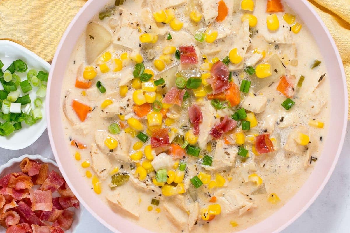 A bowl of creamy chicken and corn chowder, one of our favorite summer soups, topped with chopped green onions and bacon bits, with extra green onions and bacon served in small bowls on the side.