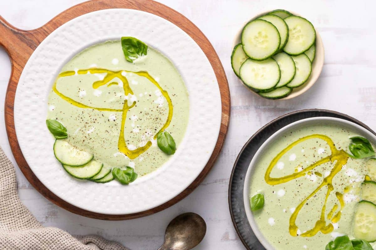 Two bowls of creamy green soup, perfect for fresh summer soups, are garnished with cucumber slices, basil leaves, a drizzle of oil, and pepper, with a small bowl of sliced cucumbers on the side.
