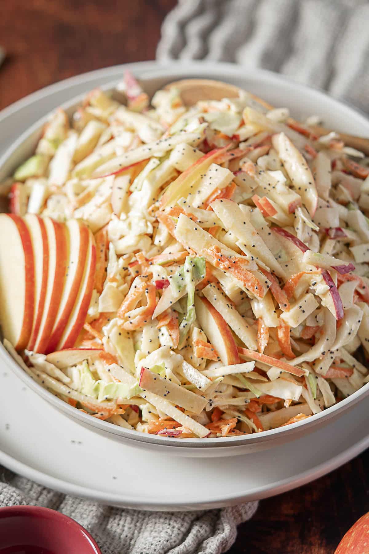 A bowl of coleslaw with shredded cabbage, carrots, and thin apple slices, served in a white dish on a wooden table.