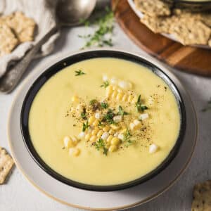 A bowl of creamy corn soup topped with corn kernels, black pepper, and fresh herbs, served on a plate with crackers nearby.