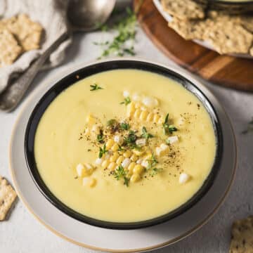 A bowl of creamy corn soup topped with corn kernels, black pepper, and fresh herbs, served on a plate with crackers nearby.