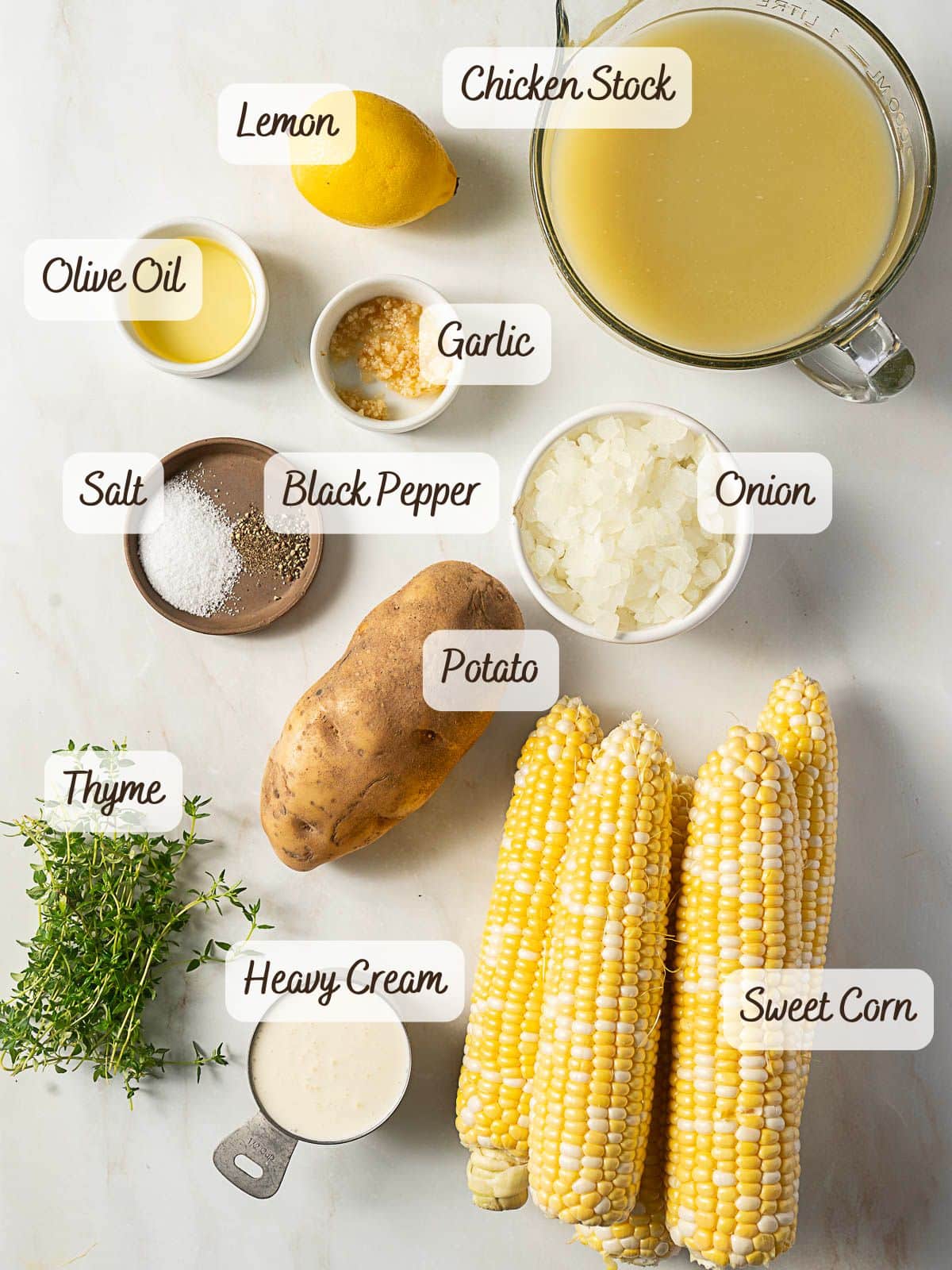 Top-down view of labeled ingredients for corn bisque or chowder: lemon, chicken stock, olive oil, garlic, salt, black pepper, onion, potato, sweet corn, thyme, and heavy cream.