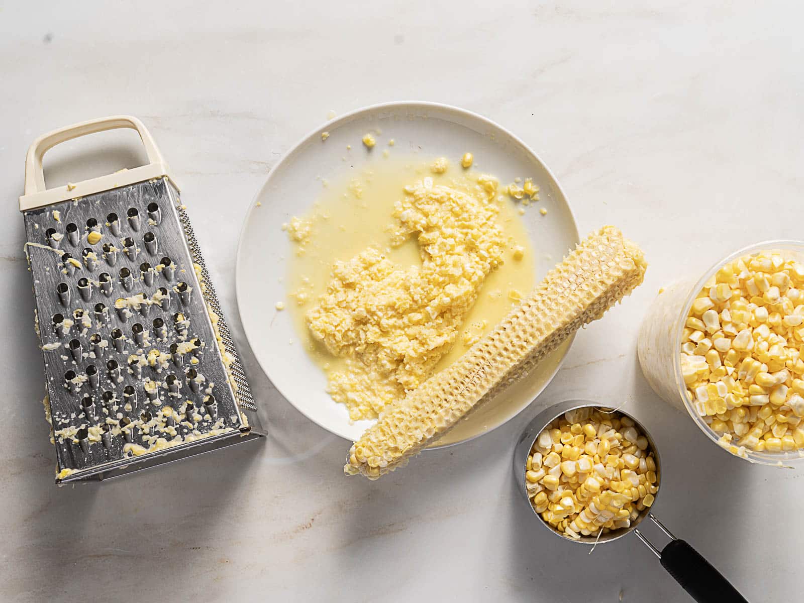 A metal grater, a plate with grated corn and a corn cob, a measuring cup, and a bowl filled with corn kernels on a white surface.