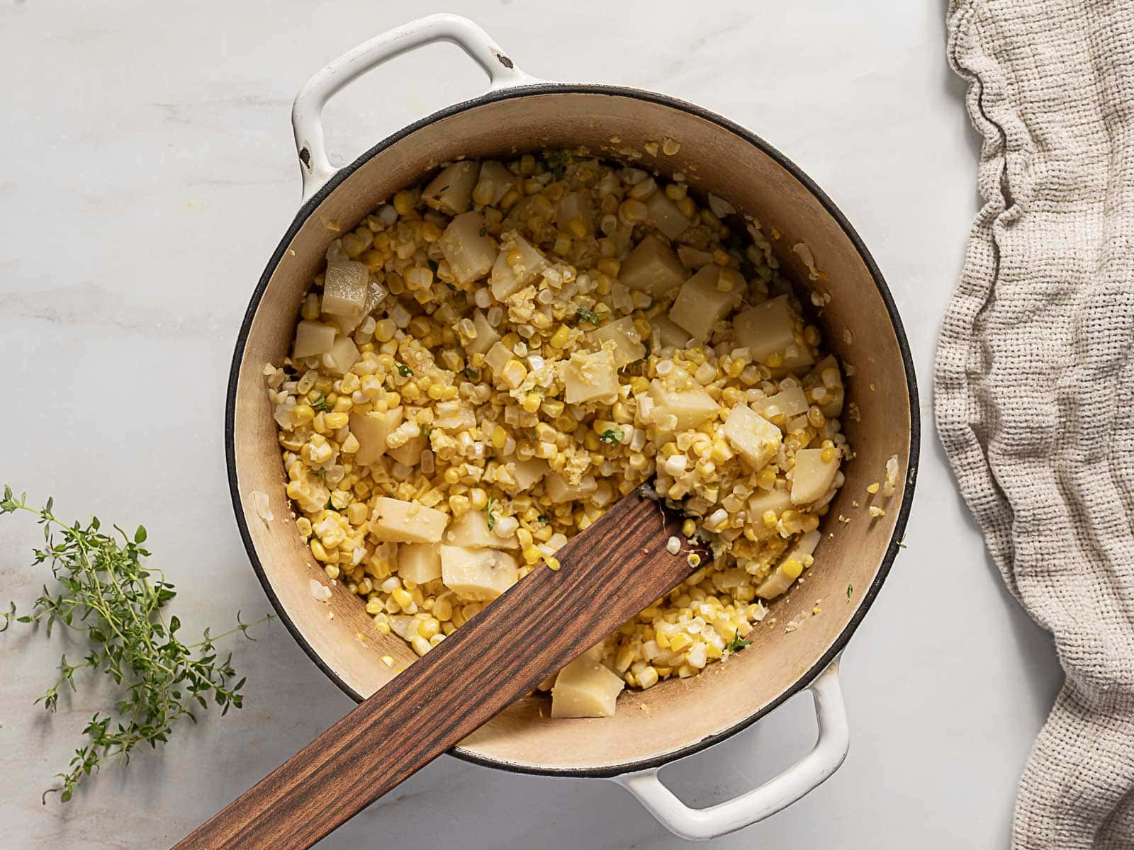 A white pot filled with a mixture of corn, diced potatoes, and herbs sits on a light surface next to a bunch of fresh thyme and a beige towel.