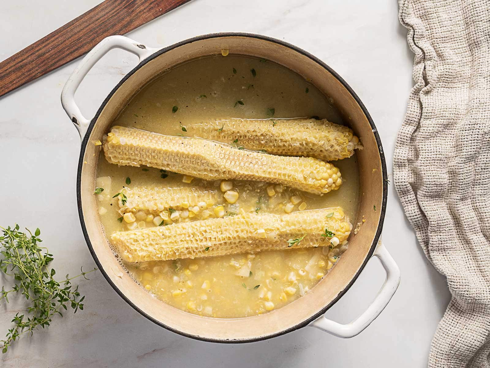 A white pot containing corn cobs simmering in a yellow broth, with corn kernels and herbs visible. A wooden board and beige cloth are nearby.