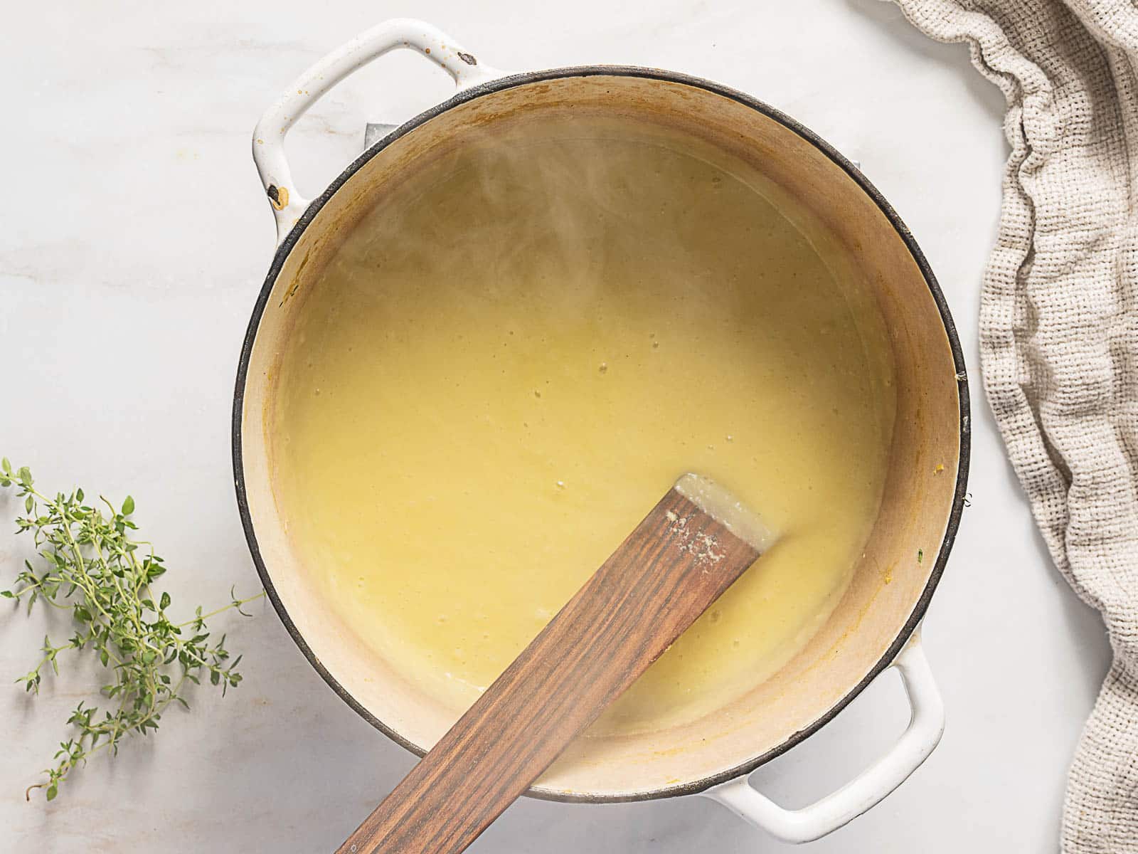 A pot of creamy yellow soup on a white countertop, with a wooden spoon inside. A sprig of fresh thyme and a beige textured cloth are nearby. Steam is rising from the soup.
