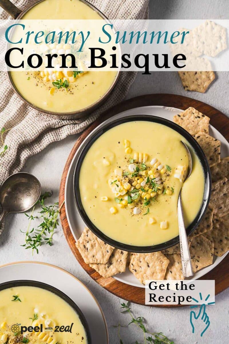 A bowl of rich corn bisque garnished with corn and herbs, served with crackers on a plate, plus a spoon and napkin nearby.