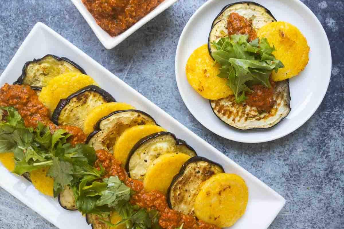 Sliced roasted Eggplant and polenta arranged on plates, topped with tomato-based sauce and fresh greens-perfect for those who love eggplant. Extra sauce served in a small bowl completes this delicious addition to your list of eggplant recipes.