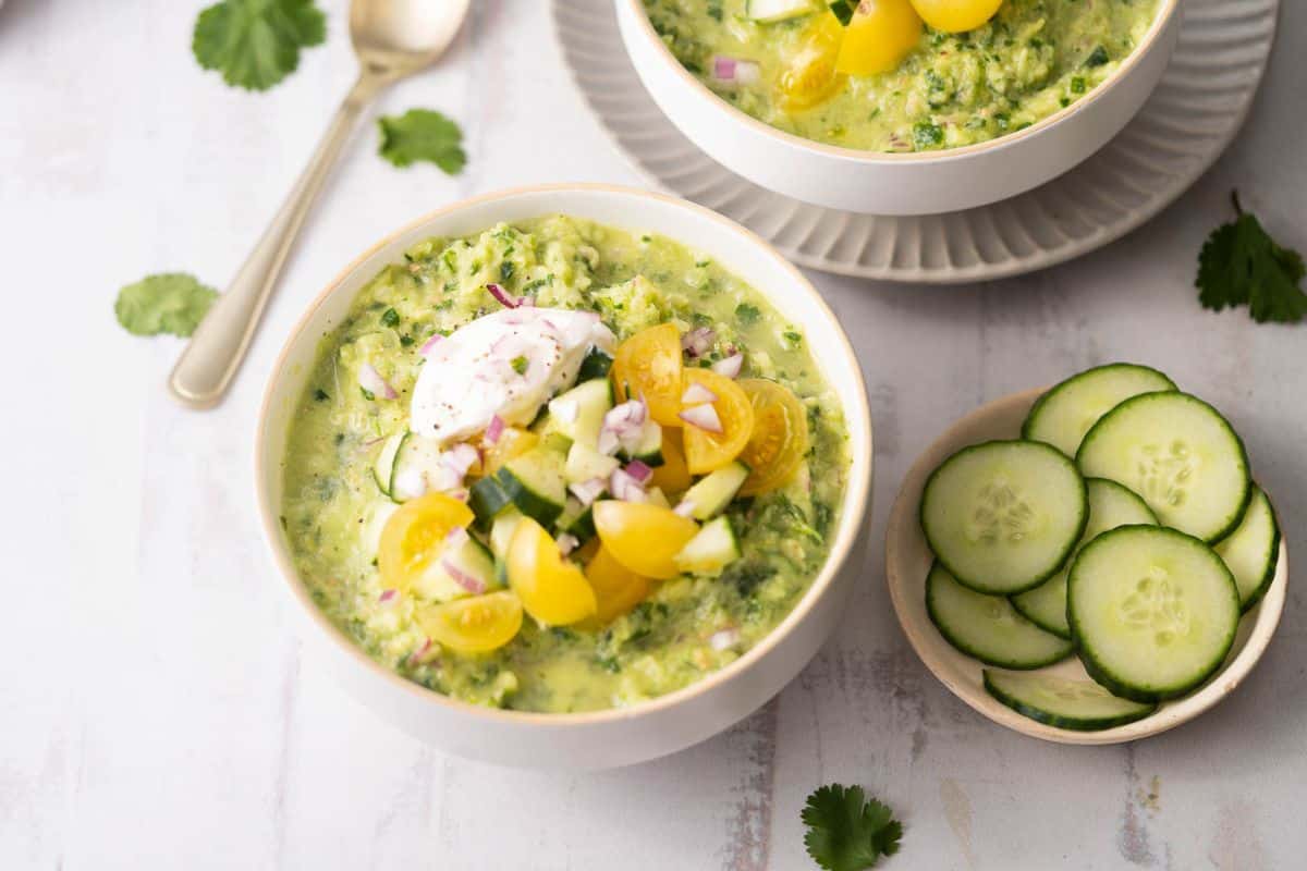 A bowl of chilled green soup, one of our favorite fresh summer soups, topped with sliced yellow tomatoes, cucumber, and red onion sits beside a small plate of cucumber slices.