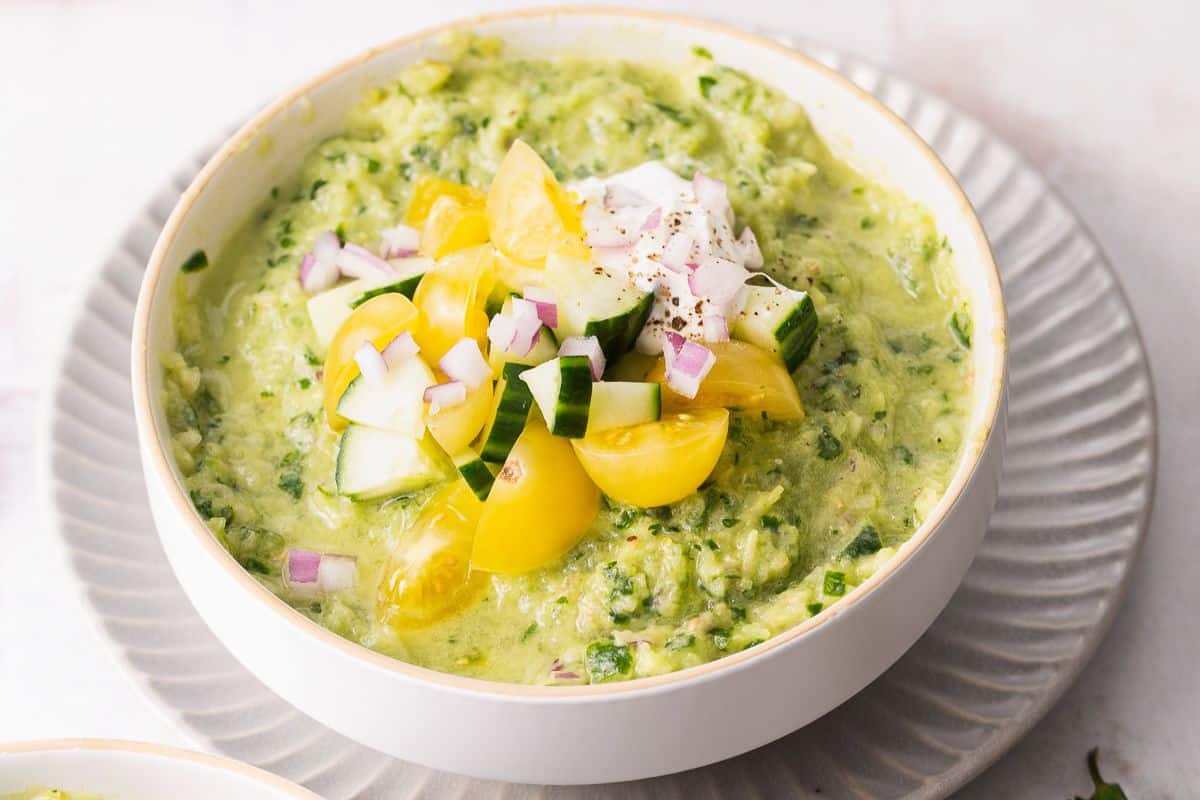 A bowl of guacamole topped with chopped yellow tomatoes, cucumber, red onion, and a dollop of sour cream, served on a plate—perfect for summer recipes and those who love fresh tomato recipes.