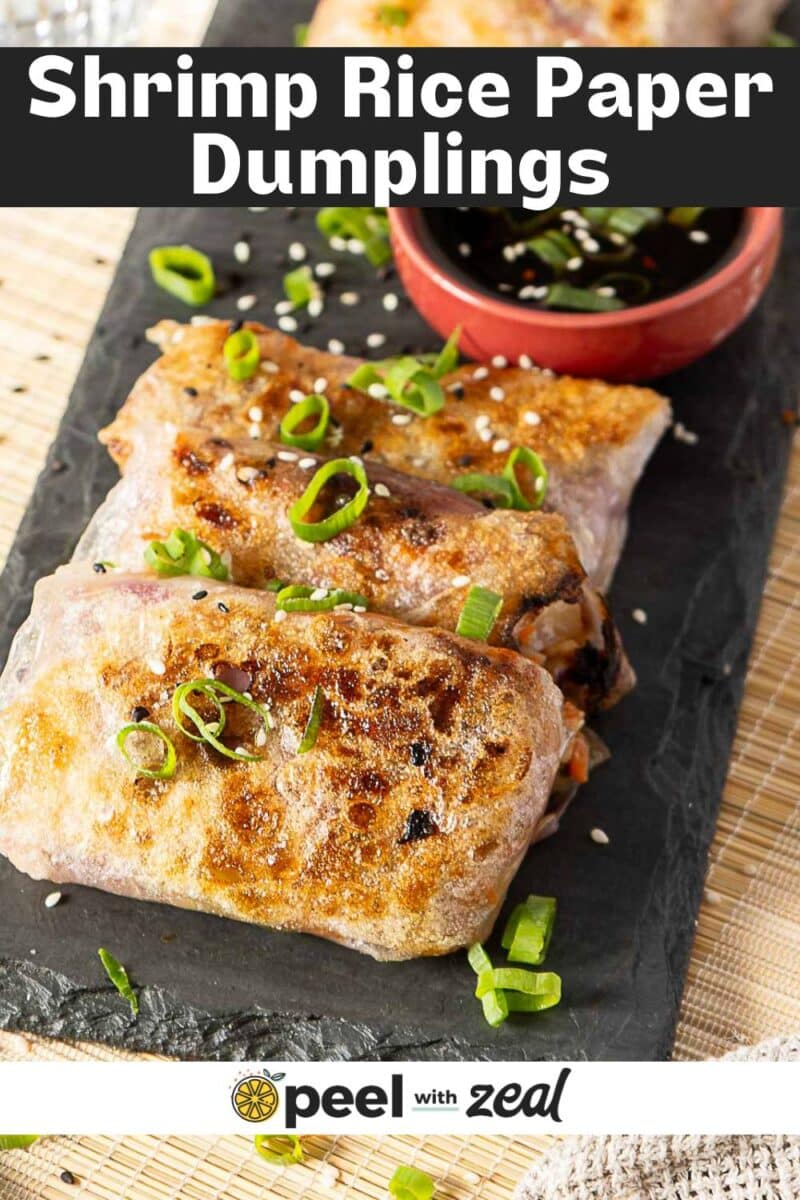 Three golden-brown rice paper dumplings filled with shrimp, garnished with sliced green onions and sesame seeds, served on a black slate alongside a small bowl of dipping sauce.
