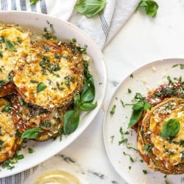 Two plates of baked eggplant parmesan, a delicious example of eggplant recipes, topped with melted cheese, tomato sauce, parsley, and basil leaves, set on a white surface with a striped napkin and utensils.