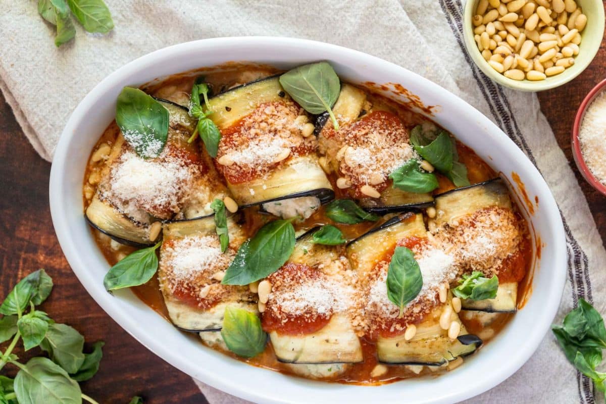 A white oval dish filled with rolled eggplant slices topped with tomato sauce, cheese, basil leaves, and pine nuts on a wooden surface-a delicious example of creative eggplant recipes.