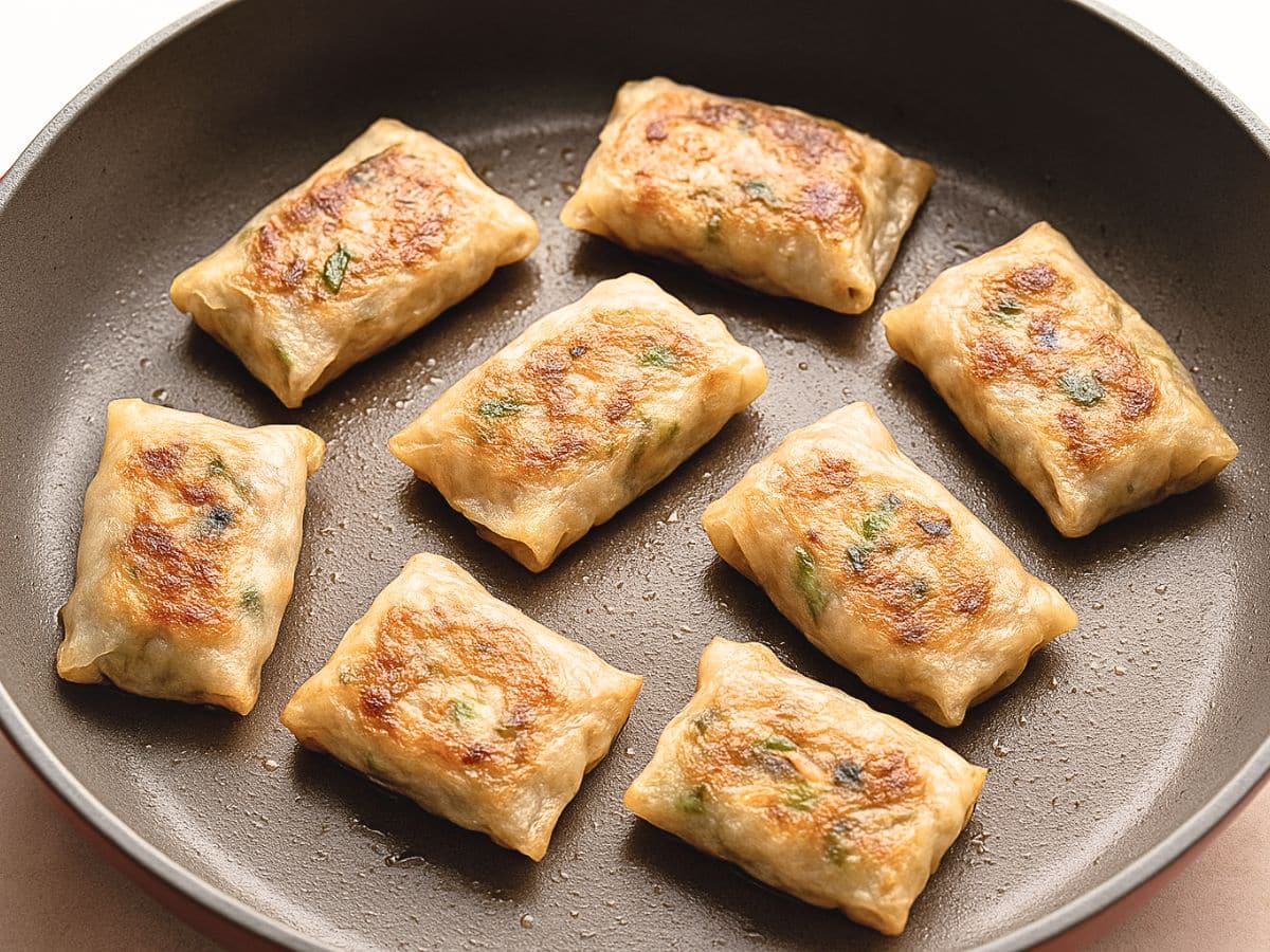 Eight rectangular dumplings are pan-frying in a skillet, showing a golden-brown, crispy surface.