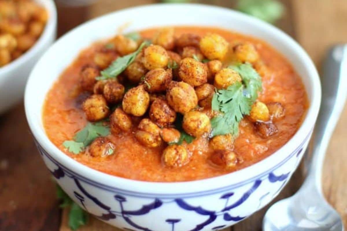 A bowl of tomato-based soup topped with roasted chickpeas and cilantro, placed on a wooden surface with a spoon nearby-perfect for those seeking healthy soups and easy summer recipes.