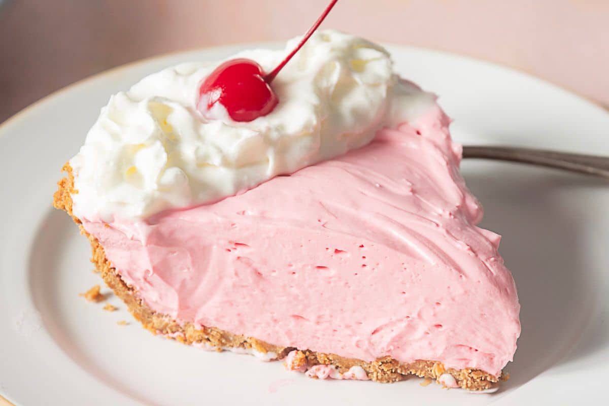 A slice of pink cream pie with a graham cracker crust, topped with whipped cream and a cherry, on a white plate with a fork.