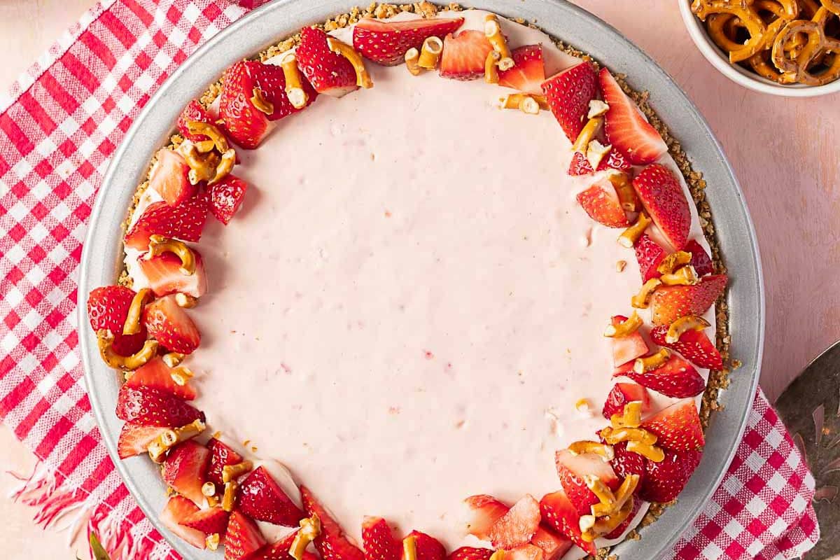 A strawberry cream pie garnished with sliced strawberries and pretzel pieces on a gray plate, set on a red and white checkered cloth.