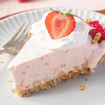 A slice of strawberry cream pie with a graham cracker crust, topped with whipped cream and a halved strawberry, on a plate with a fork.