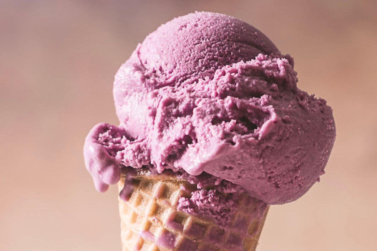 A close-up of a waffle cone holding two scoops of purple ice cream against a blurred background.