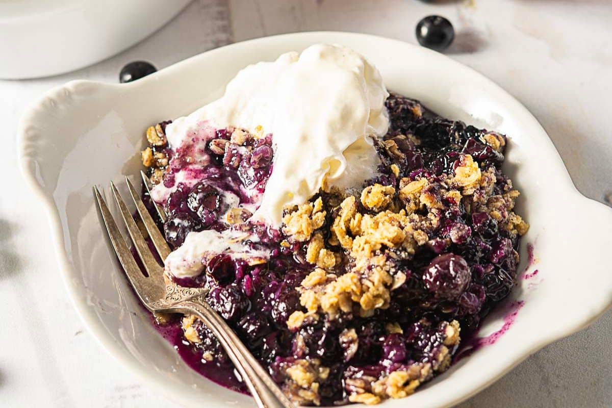 A bowl of blueberry crisp topped with a scoop of vanilla ice cream, served with a fork on a white plate.