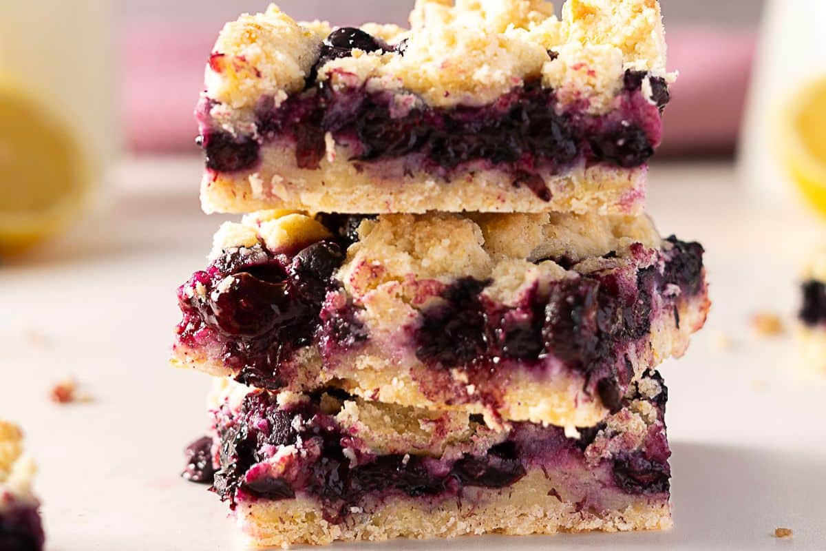 A stack of three blueberry crumb bars with a crumbly topping and layers of blueberry filling and crust on a light surface.