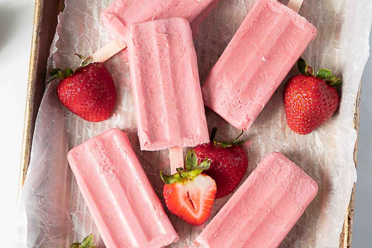 Pink strawberry popsicles arranged on parchment paper in a tray, with whole and halved fresh strawberries placed around them.