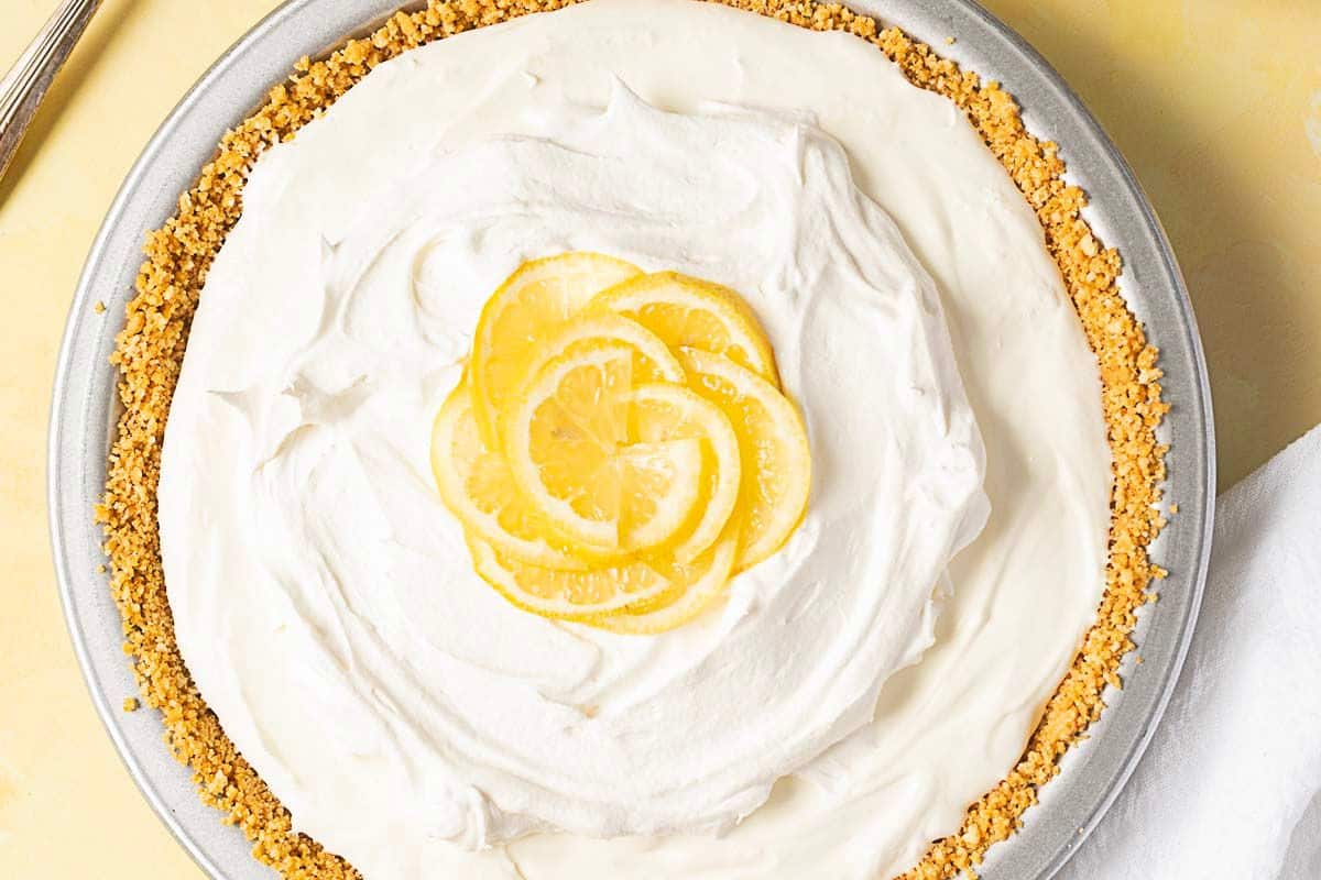 A pie with a crumb crust, topped with a layer of whipped cream and garnished with thin lemon slices in the center.