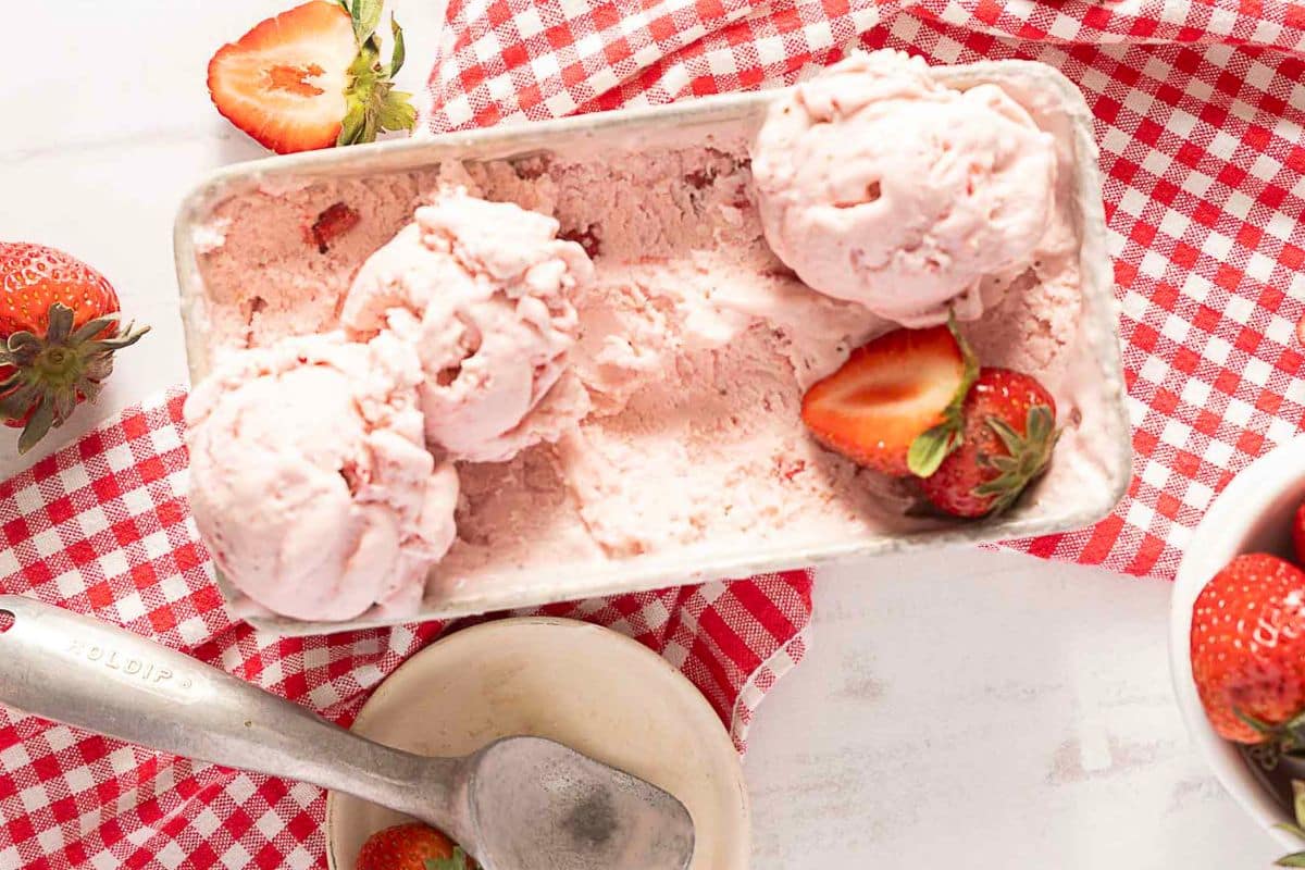 Rectangular container with scoops of strawberry ice cream and fresh strawberries, on a red and white checkered cloth, with an ice cream scoop and a small bowl nearby.