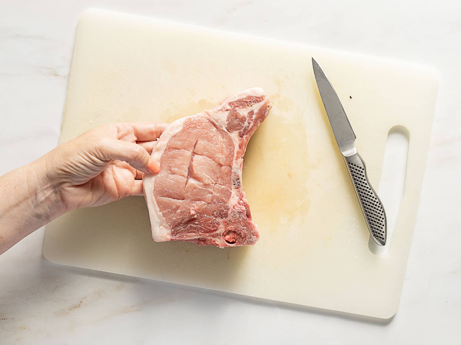 A hand holds a raw pork chop on a white cutting board next to a paring knife.