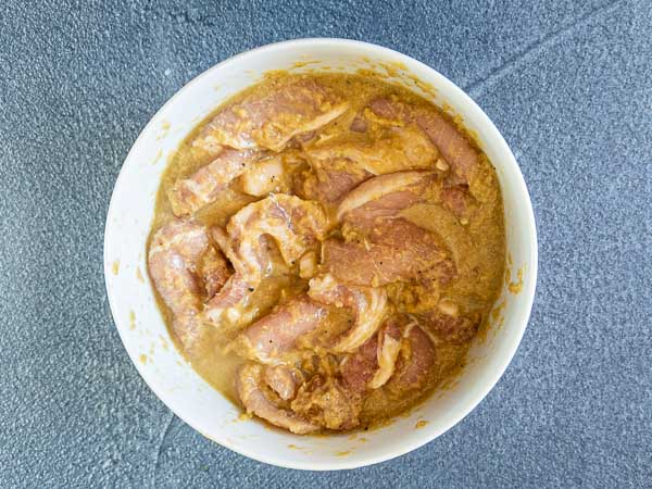 Raw chicken pieces marinating in a yellowish spice mixture in a white bowl on a gray surface.