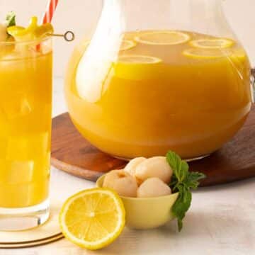 A glass and pitcher of yellow lychee lemonade with ice and lemon slices, garnished with lychee and mint, on a wooden board with a cut lemon and a small bowl of lychees—perfect for those seeking refreshing drinks or non alcoholic summer beverages.