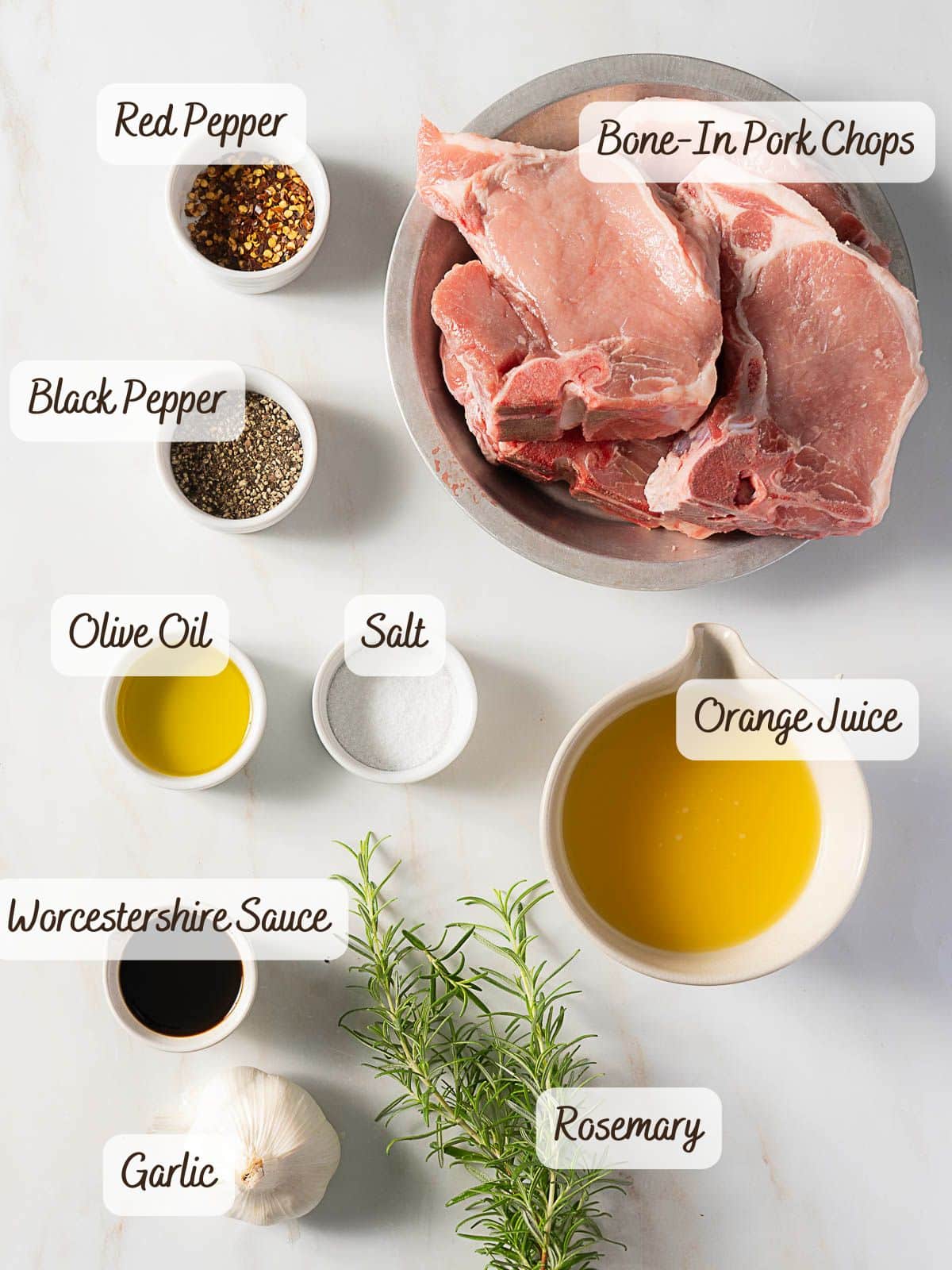 Flat lay of labeled ingredients for marinated grilled pork chops, including bone-in pork chops, orange juice, Worcestershire sauce, olive oil, salt, black pepper, red pepper, garlic, and fresh rosemary on a white surface.