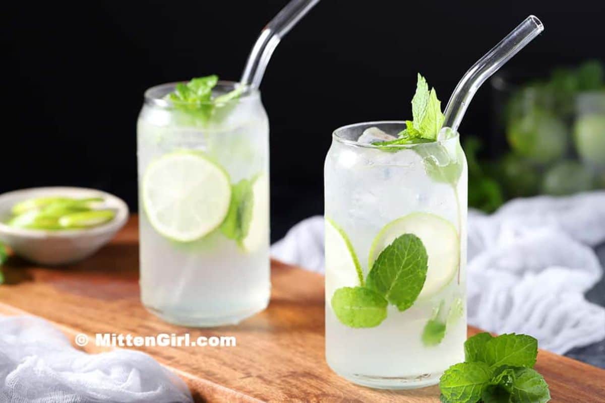 Two clear glasses with glass straws, filled with a lime and mint drink on a wooden board, garnished with fresh mint and lime slices-perfect refreshing drinks to enjoy as non alcoholic drinks this summer.