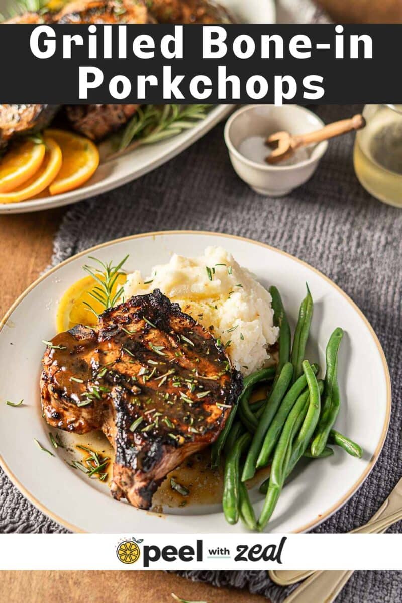 A plate with a marinated grilled pork chop, green beans, mashed potatoes, and orange slices sits on a gray placemat with a fork and napkin nearby.
