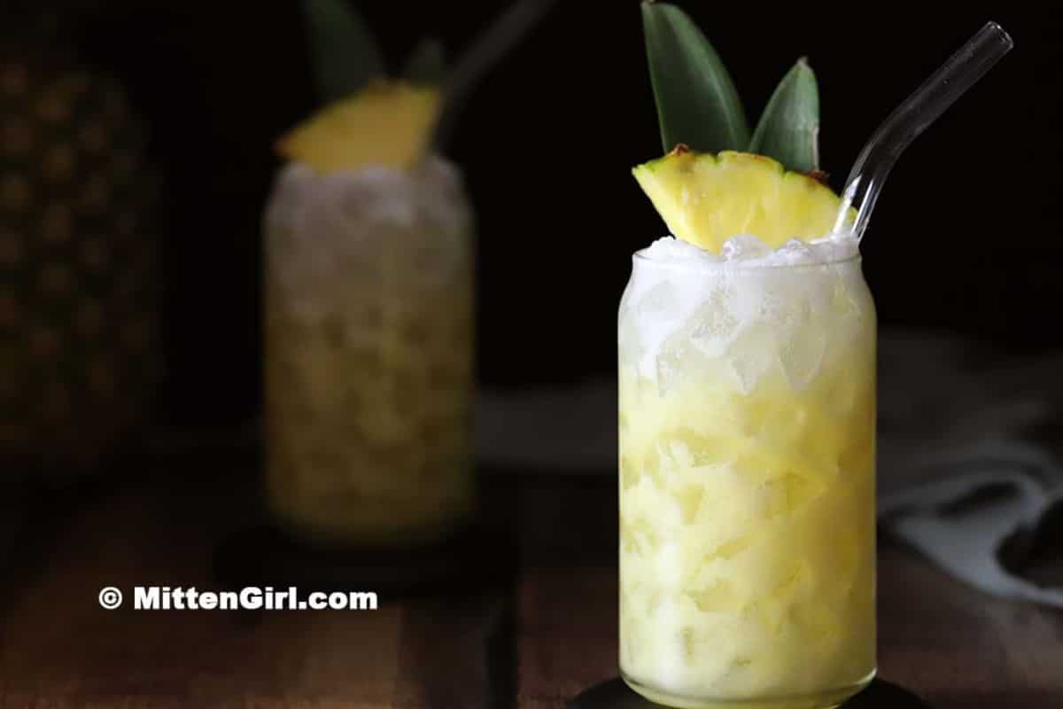 A glass filled with a yellow iced drink, garnished with a pineapple slice and two pineapple leaves, with a clear straw-perfect for those seeking refreshing drinks. Another similar summer beverage is blurred in the background.