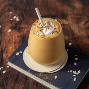 A glass of pumpkin smoothie with whipped cream, oats, and cinnamon on top, served with a straw on a coaster and book, with oats scattered on a wooden surface.