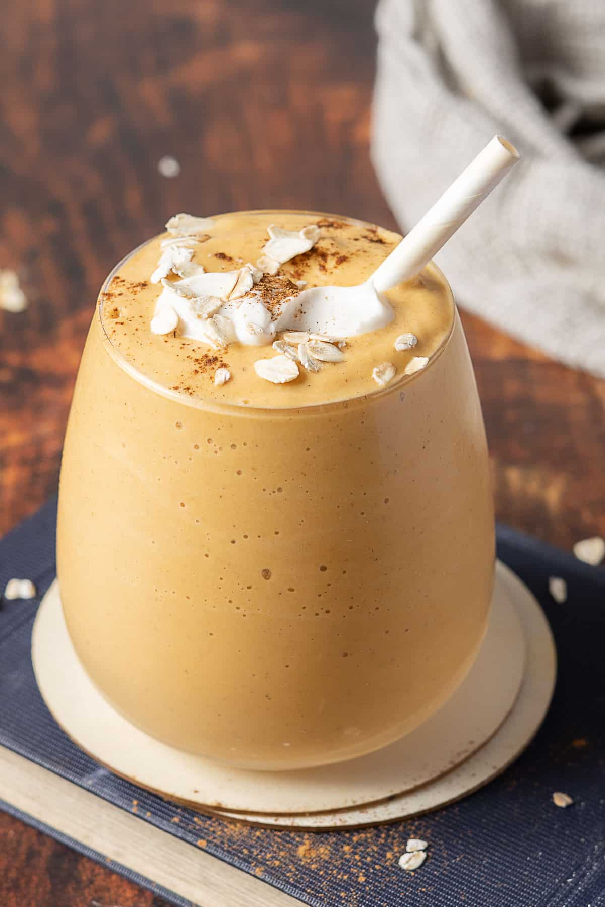 A glass of thick orange smoothie topped with oats and cinnamon sits on a coaster, with a white spoon sticking out.
