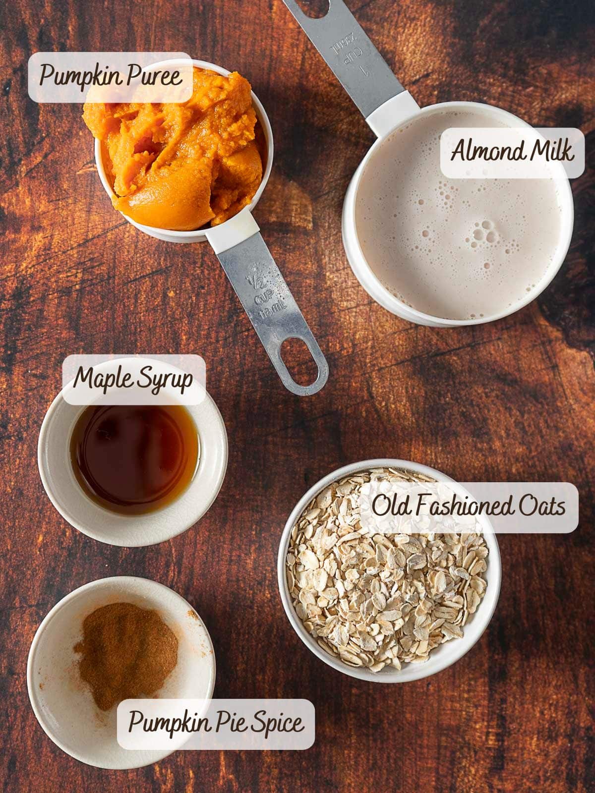 Five labeled ingredients in bowls and measuring cups—pumpkin puree, almond milk, old fashioned oats, maple syrup, and pumpkin pie spice—arranged on a wooden surface, perfect for blending into a creamy pumpkin smoothie.