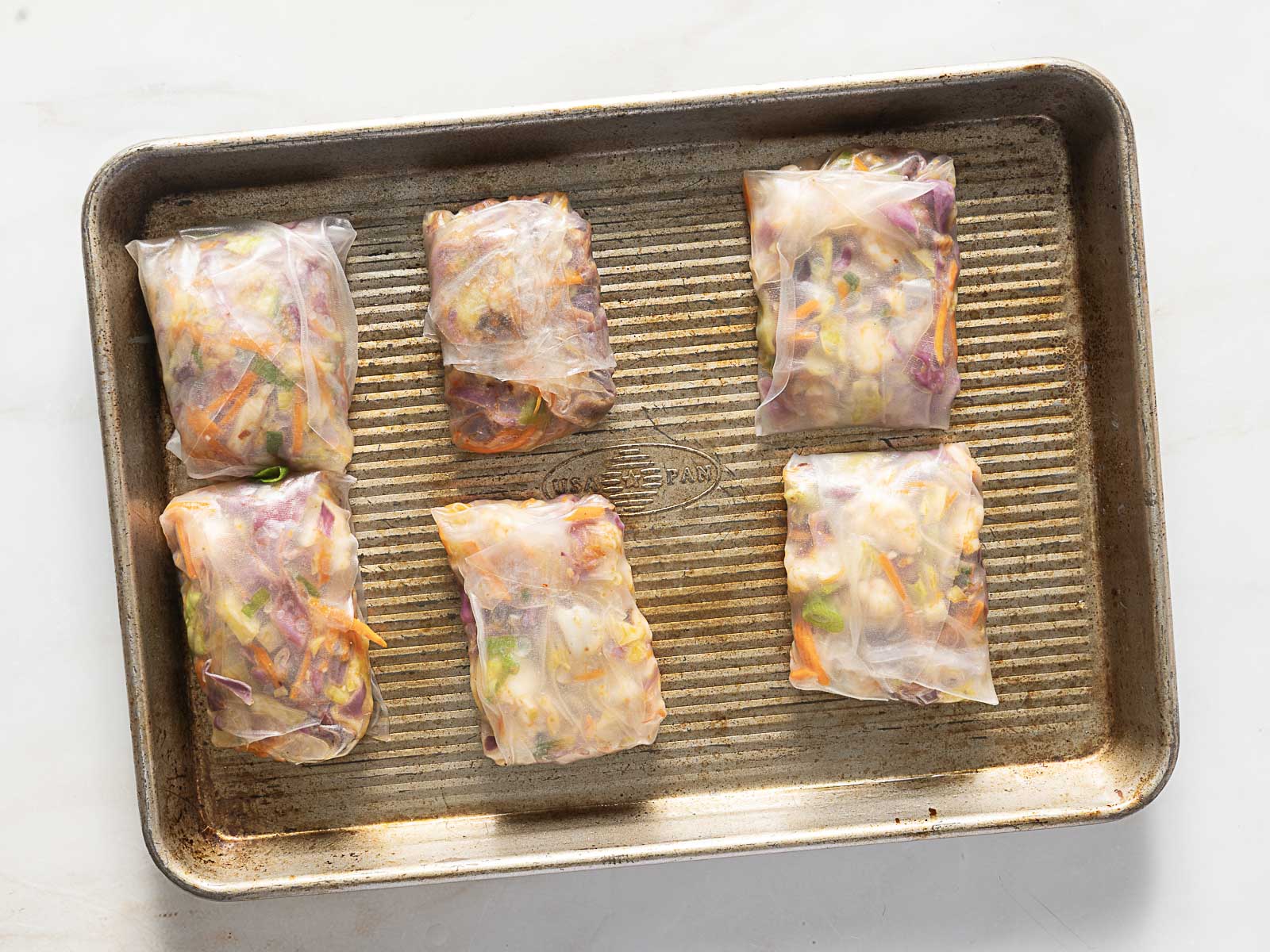 Six rectangular spring rolls with visible colorful vegetable filling are arranged on a metal baking sheet.