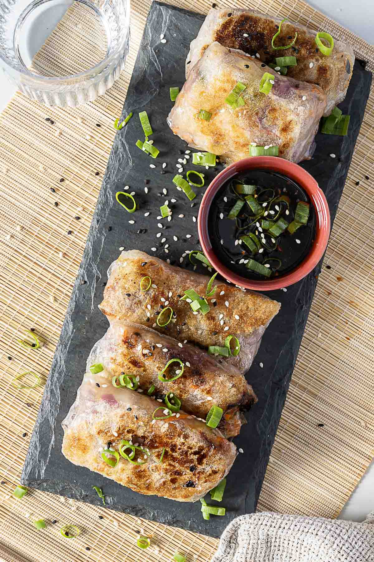 Four browned spring rolls garnished with chopped green onions and sesame seeds, served on a black slate with a small bowl of soy sauce and a glass of water on the side.