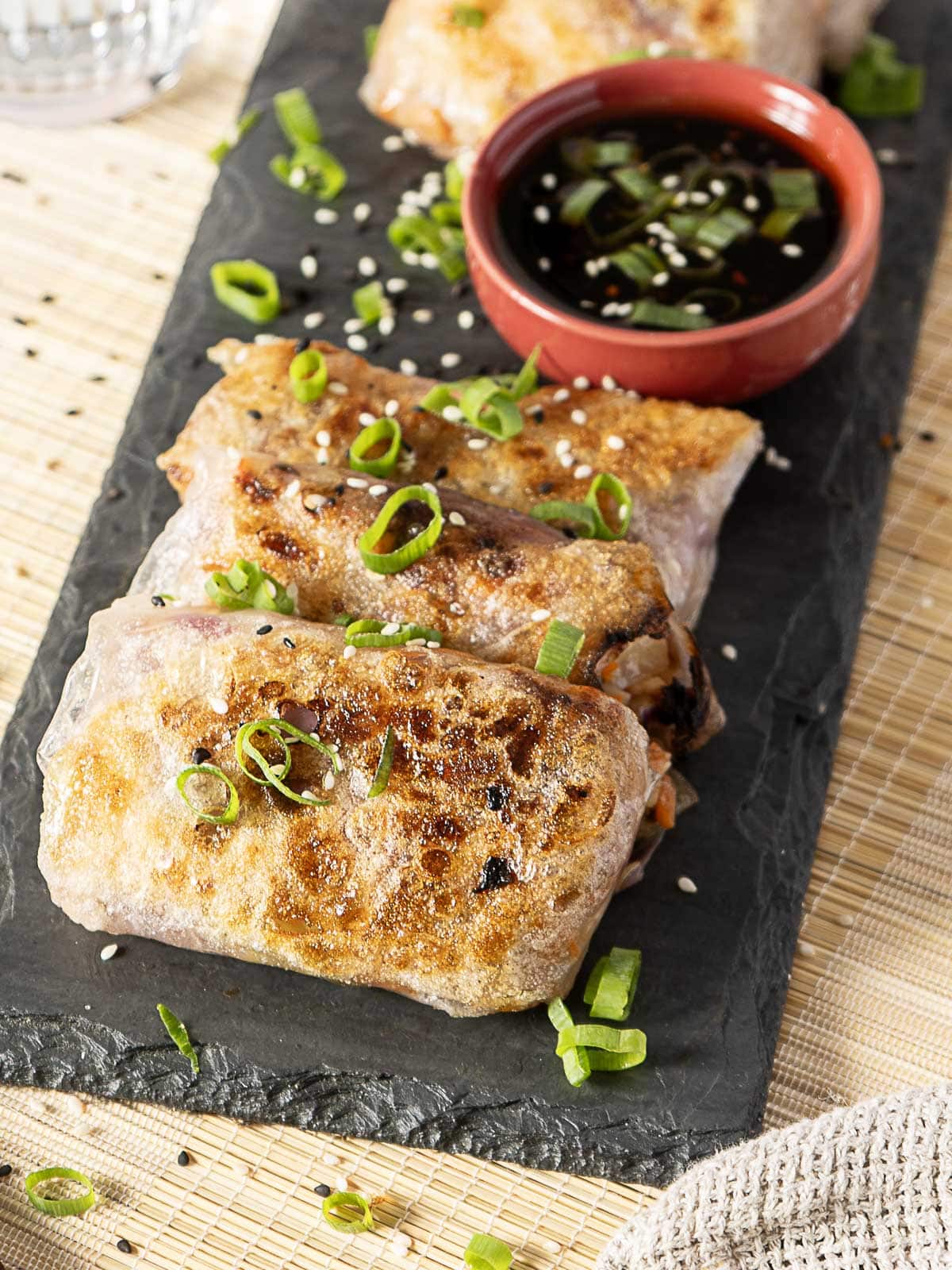 Three crispy spring rolls garnished with chopped green onions are served on a black slate plate next to a small red bowl of soy dipping sauce.