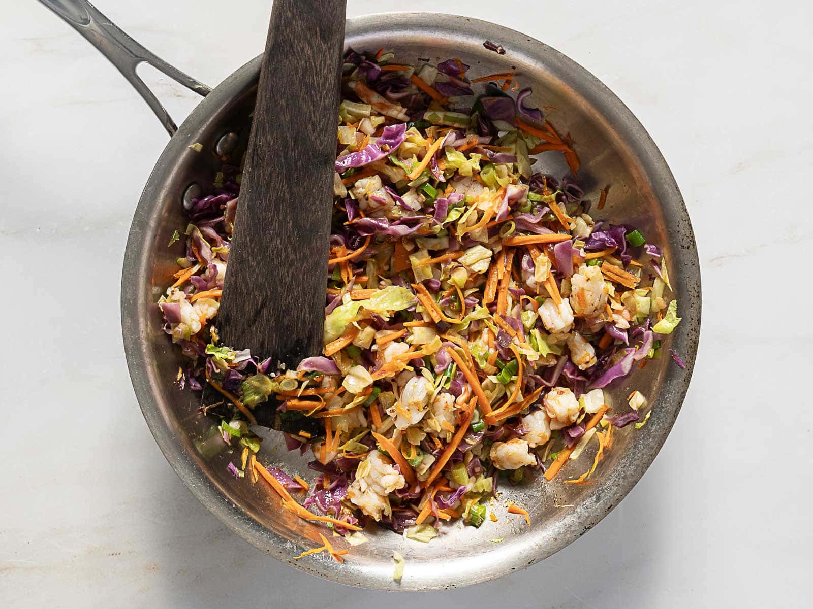 A stainless steel pan contains a mixture of shredded cabbage, carrots, and pieces of chicken being stirred with a wooden spatula.