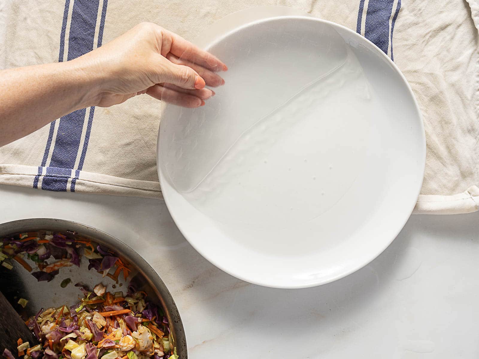A hand dips a round sheet of rice paper into a shallow white plate filled with water; a skillet of cooked vegetables sits nearby.
