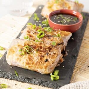 Three pan-fried spring rolls garnished with chopped green onions are served on a slate tray with a small bowl of dipping sauce.
